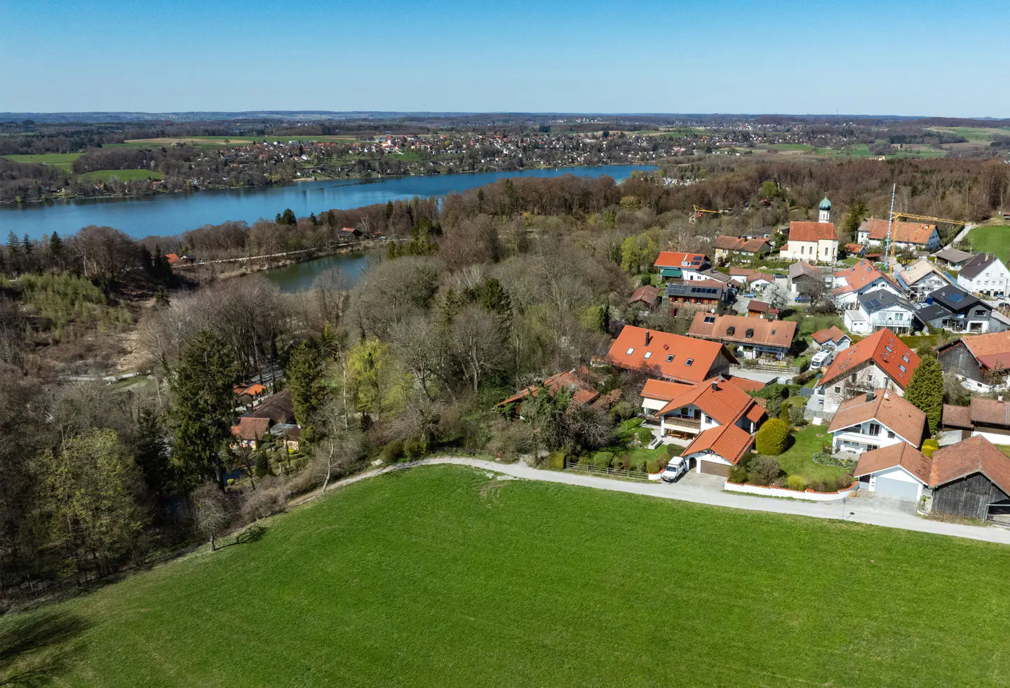 Aerial view of a village with red-roofed houses, a church, and a lake in the background under a clear blue sky.