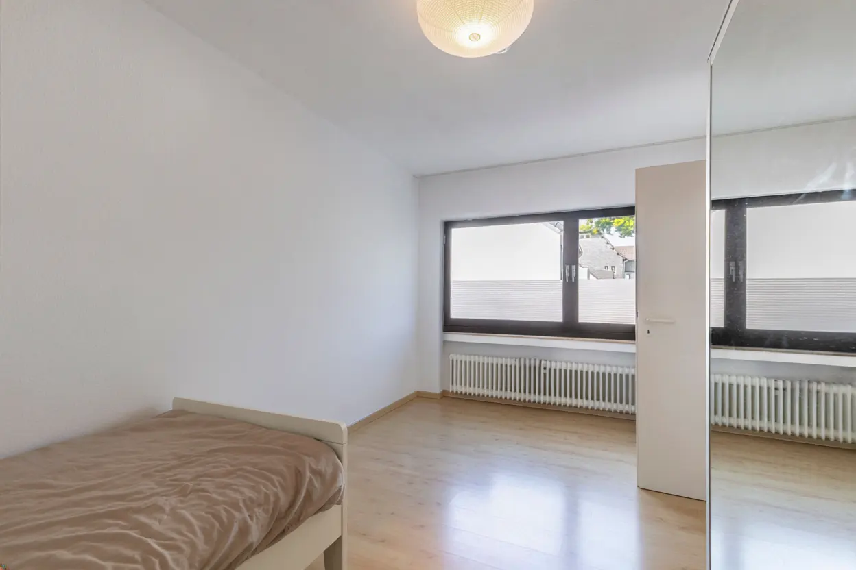 Bright bedroom with white walls, wood floor, and a single bed with brown bedding. A window with blinds and a radiator are visible. A mirrored closet door reflects the room.