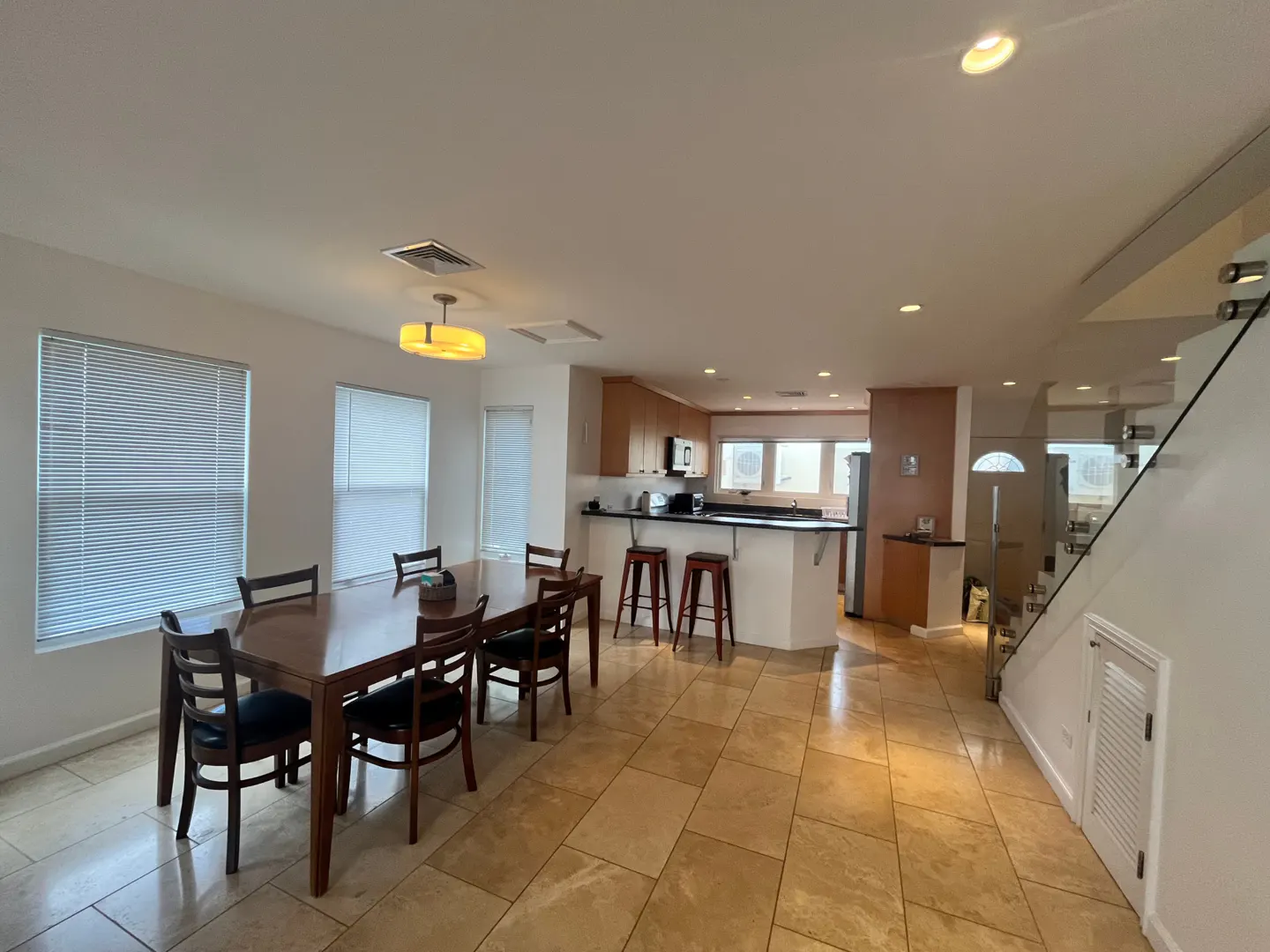 Open-concept home interior with dining table, kitchen island with stools, and staircase with glass railing. Tiled floor.