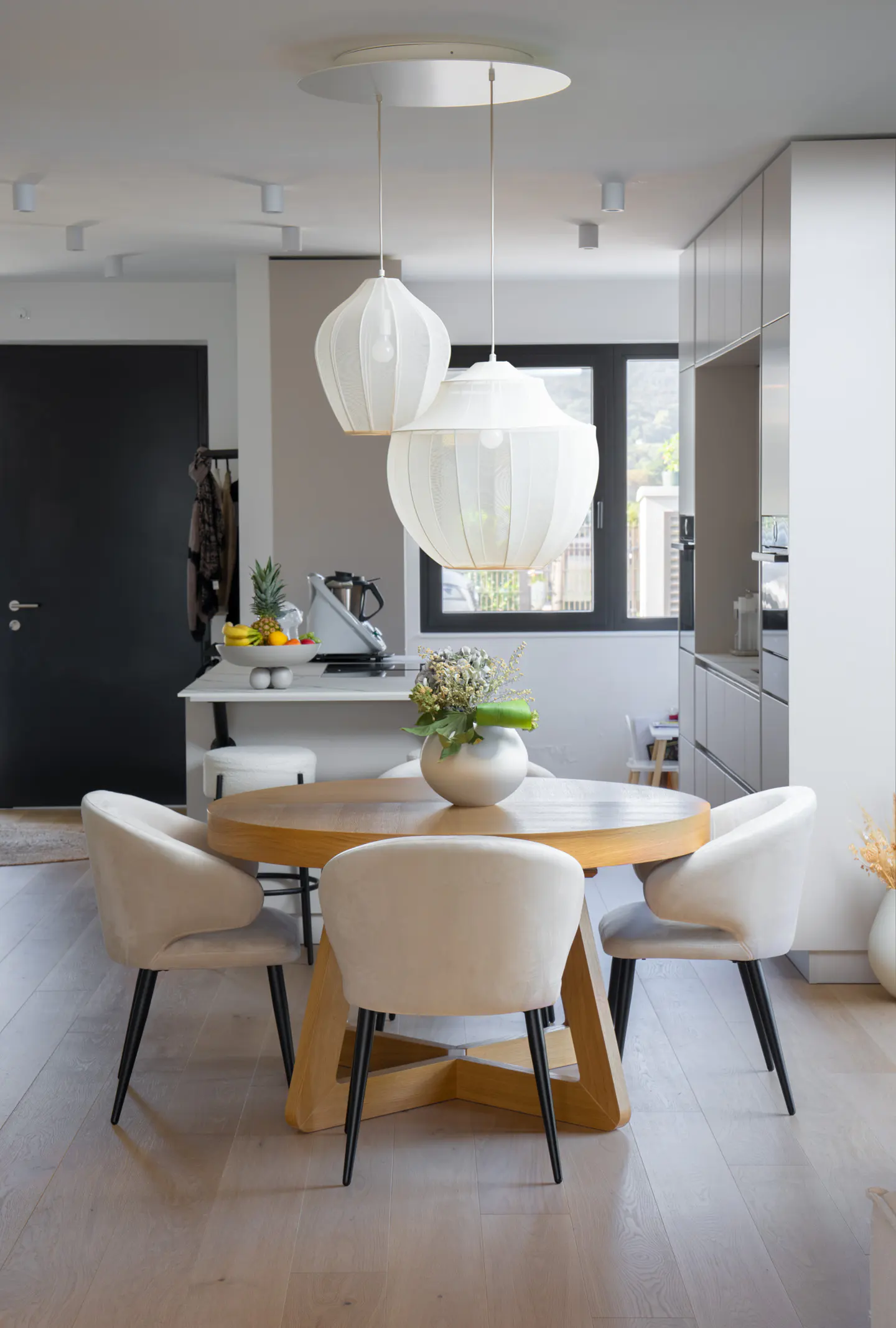 Bright, modern dining area with a round wooden table, four cream-colored chairs, and two white pendant lights. A vase of flowers sits on the table.