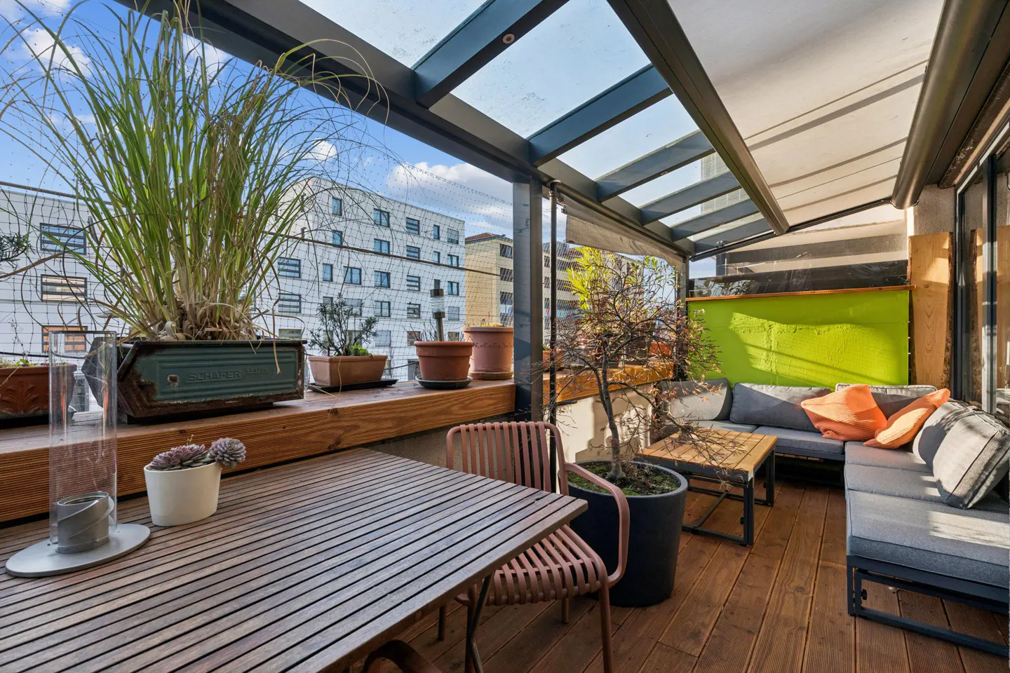 A covered balcony with a wooden floor features a table, chairs, and a gray sofa with orange pillows. Plants and city views add to the outdoor living space.