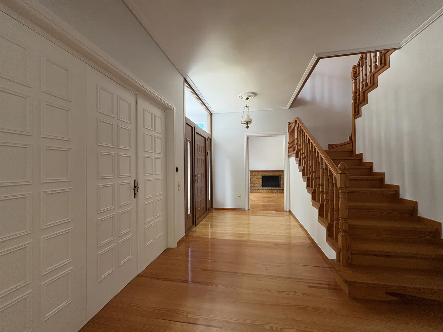 Bright foyer with hardwood floors, a wooden staircase, white paneled doors, and a view into a room with a brick fireplace.