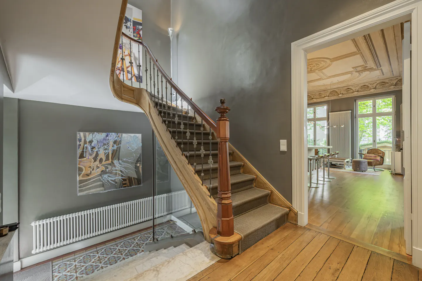Interior view of a home featuring a wooden staircase with a carpet runner, gray walls, and a glimpse into a living room with large windows.