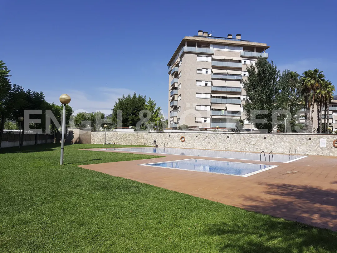 Outdoor swimming pool with blue water, surrounded by a green lawn and a paved deck, with a tall apartment building behind.