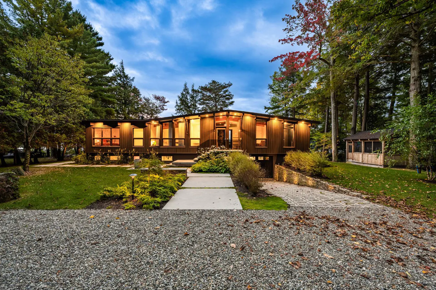 Exterior view of a modern brown house with large windows, a stone walkway, and lush green landscaping.