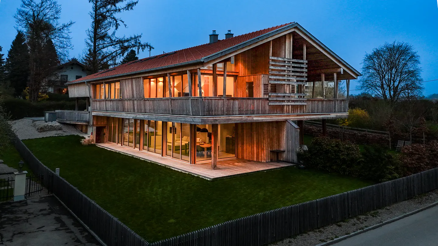 Two-story wood house with a red tile roof, large windows, and a green lawn at dusk.