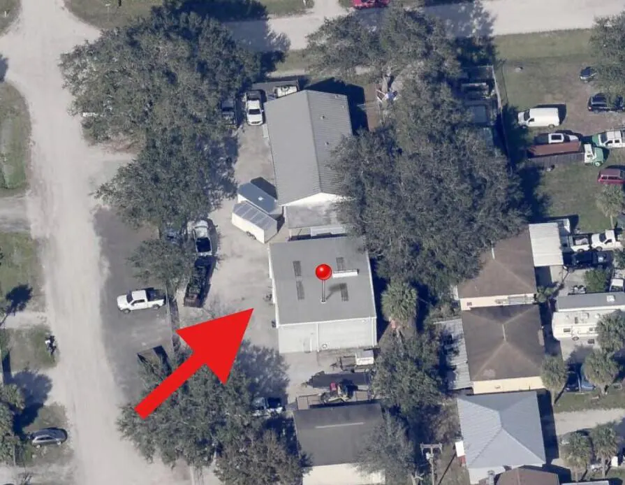 Aerial view of a property with a gray building marked by a red pin, surrounded by trees and parked vehicles. A red arrow points towards the building.