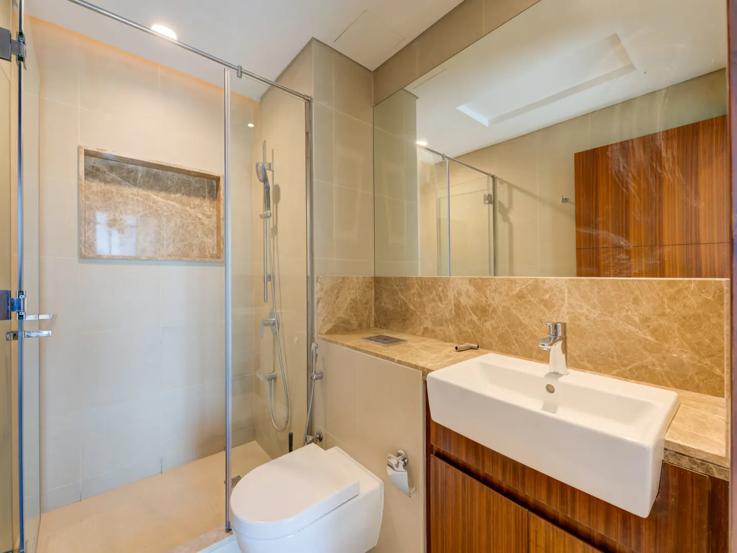 Modern bathroom with glass shower, white toilet, and rectangular sink with wood cabinet. Marble accents and a large mirror add elegance.