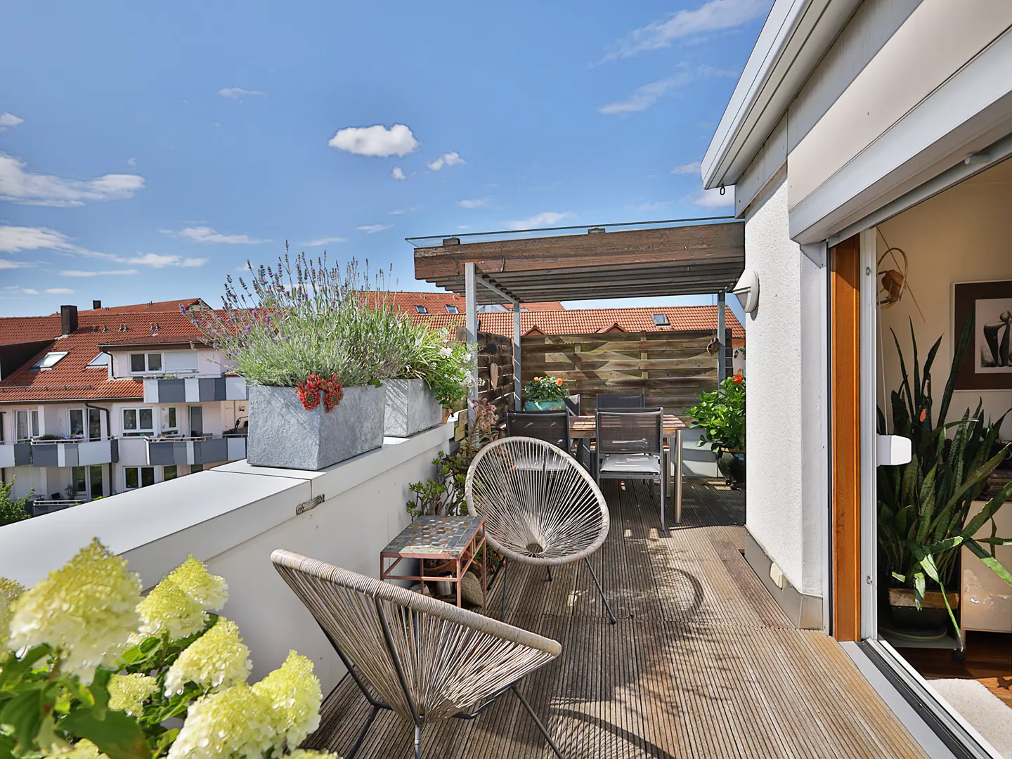 A sunny balcony with wooden floors, chairs, and potted plants. A table and chairs are under a pergola. Buildings are visible in the background.
