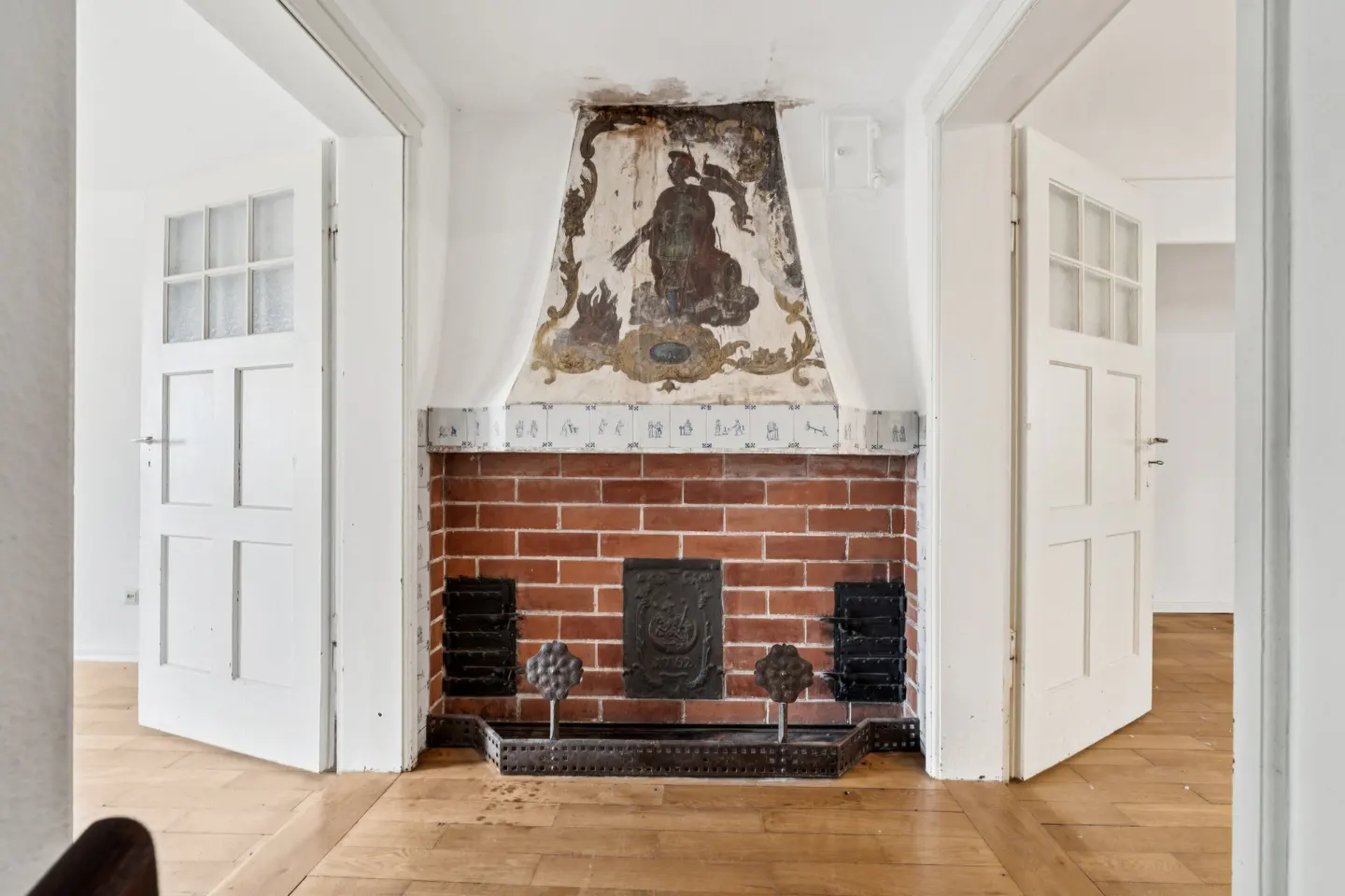 Interior view of a brick fireplace with a mural above, flanked by open white doors on a wood floor.