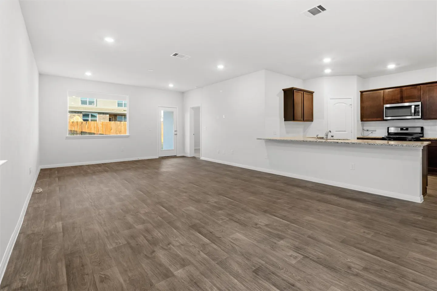 An open-concept living space with wood-look floors, white walls, and dark wood kitchen cabinets. A window and glass door provide natural light.