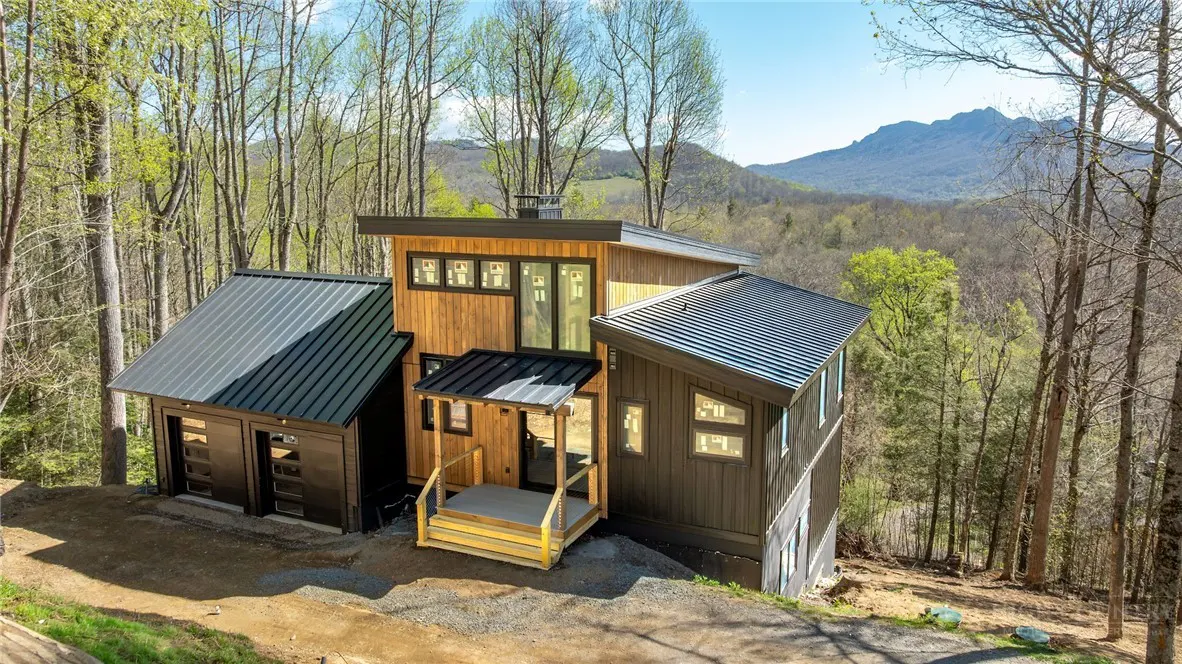 Modern wood home with black metal roof, garage, and porch, nestled in a forest with mountains in the background.