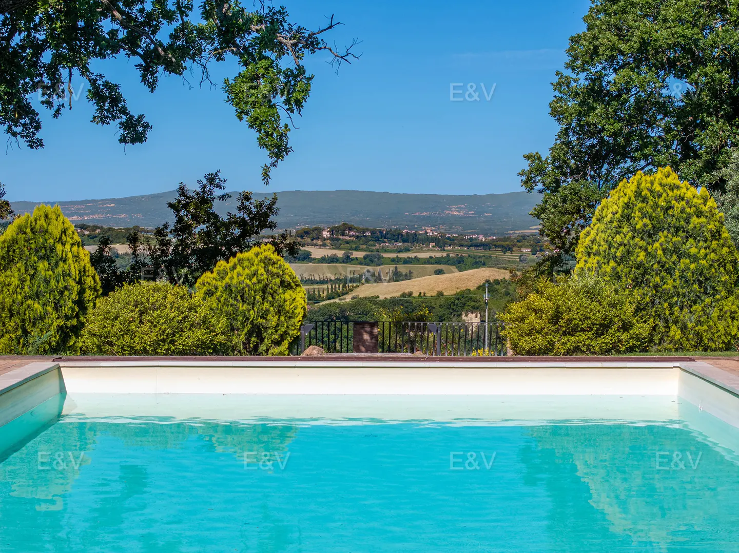 A turquoise pool overlooks a green, hilly landscape under a clear blue sky. Trees frame the top of the image.