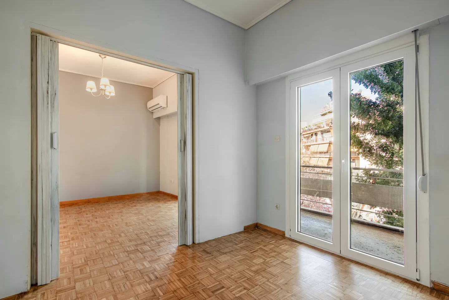 Empty room with parquet flooring, gray walls, and white trim. A doorway leads to another room with a chandelier and AC unit. Balcony doors show trees and buildings.
