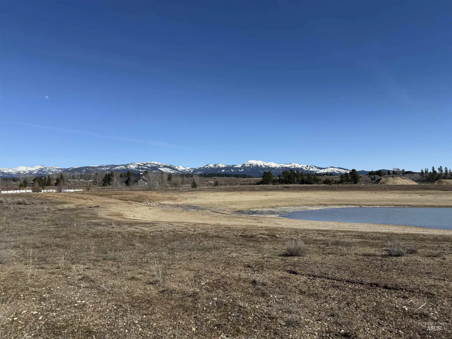 A landscape view of a dry field with a small pond, snow-capped mountains, and a clear blue sky.