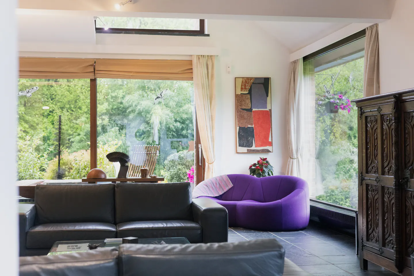 Bright living room with black leather sofas, purple couch, and large windows overlooking a lush green garden. Abstract art hangs on the wall.