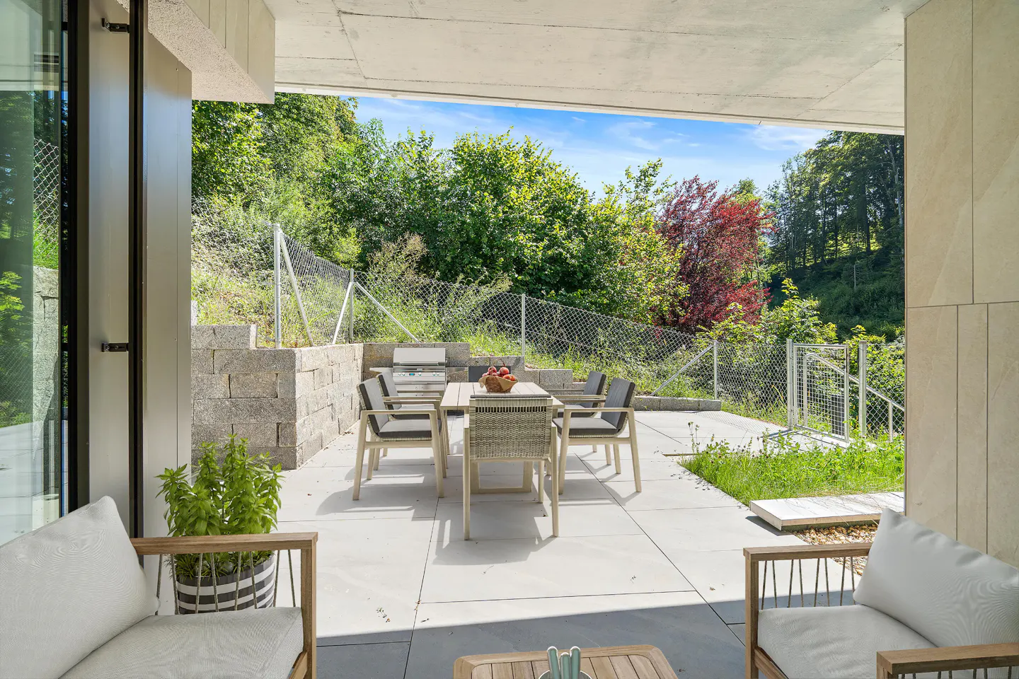Modern patio with dining set, outdoor grill, and lounge chairs overlooking a lush green backyard with a chain-link fence.
