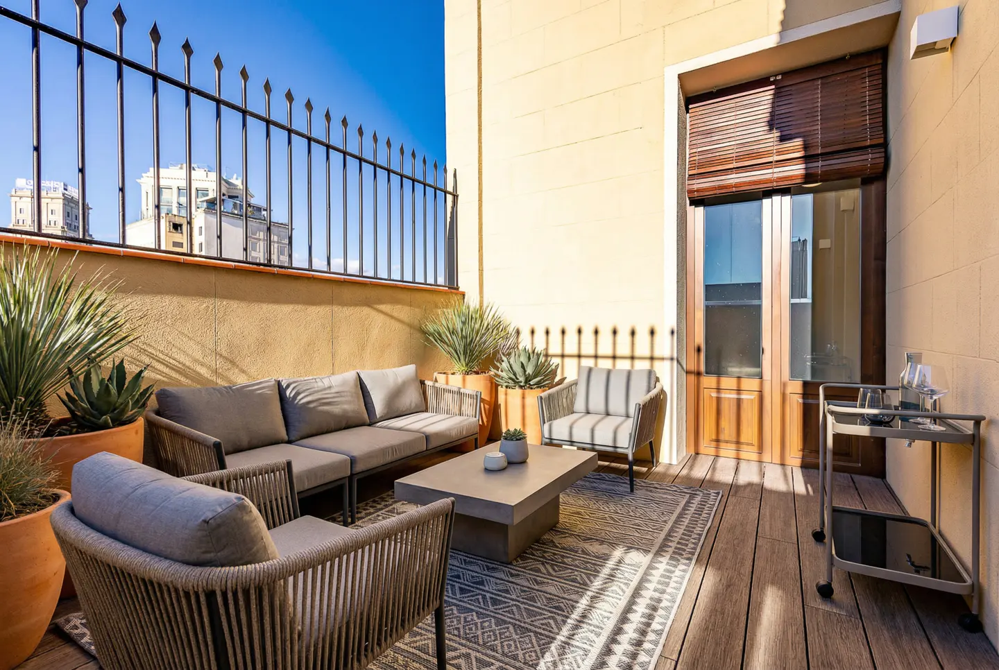 Outdoor patio with gray sofa, two chairs, and a concrete coffee table on a patterned rug. Plants in terracotta pots and a bar cart are also present.