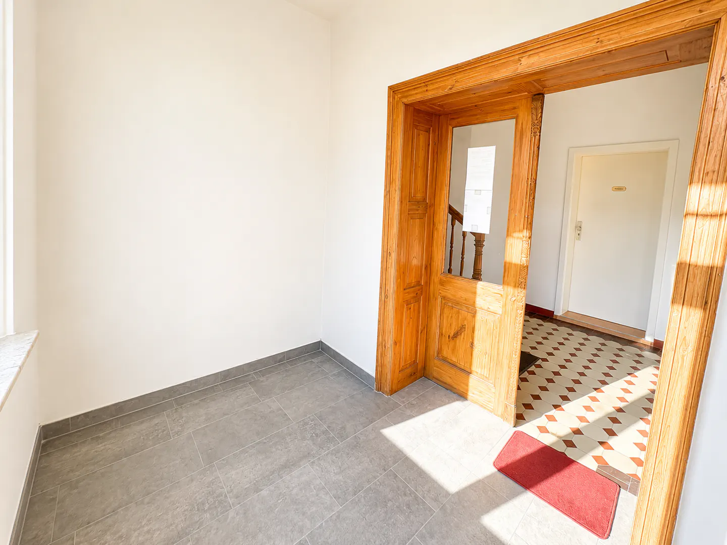 A bright entryway with grey tiled floors leading through a wooden door frame to a hallway with patterned tile flooring.