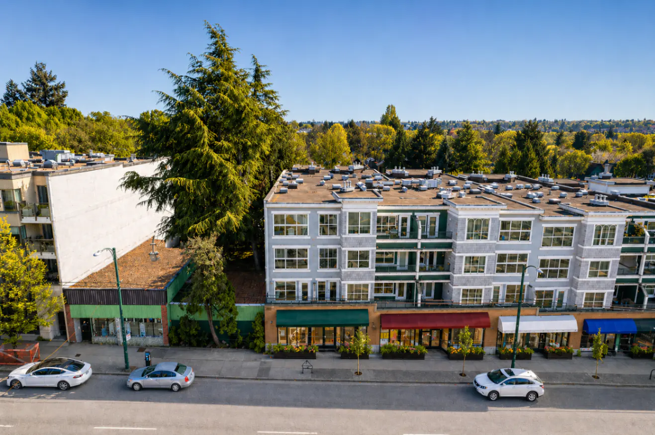 Aerial view of a gray apartment building with green and red awnings over storefronts on a tree-lined street. Cars are parked along the curb.