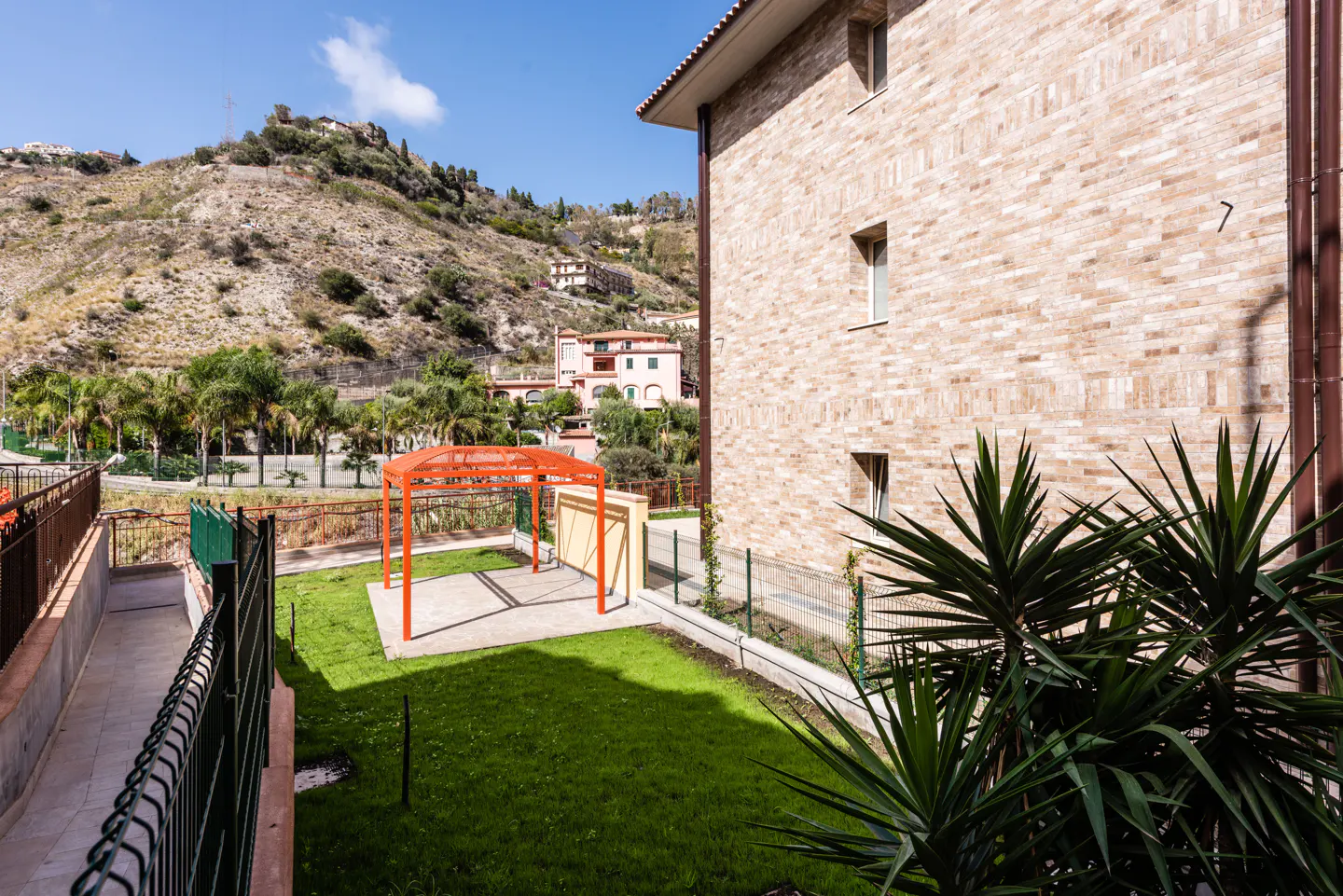 A tan brick building overlooks a green lawn with an orange metal pergola, set against a backdrop of a sunny hillside.
