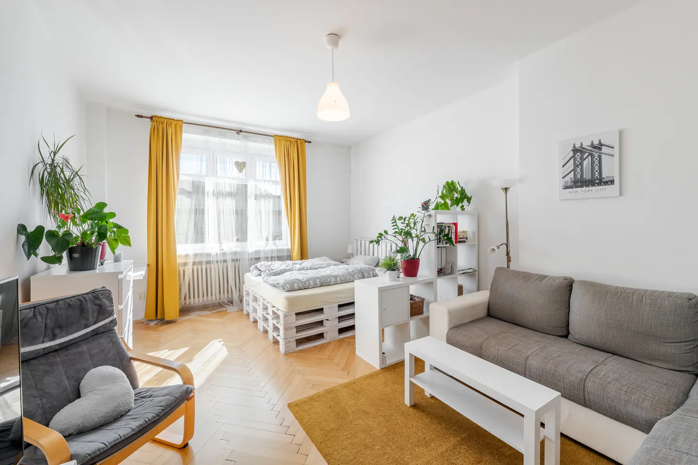 Bright studio apartment with a pallet bed, grey sofa, yellow curtains, and hardwood floors.