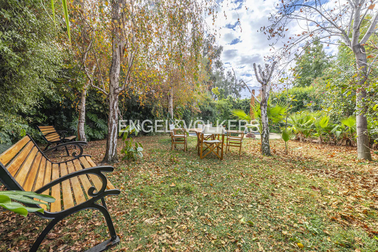 Outdoor patio with wooden table and chairs, benches, trees, and fallen leaves on the ground. Engel & Völkers logo in the background.
