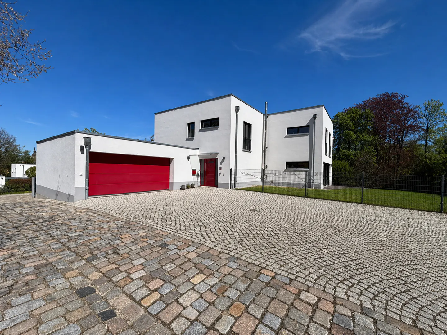 Modern white house with a red garage door and cobblestone driveway under a clear blue sky.