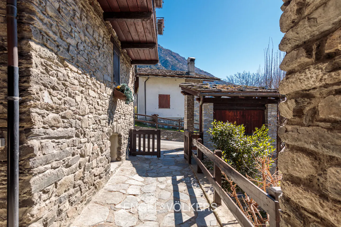 Stone pathway between buildings leads to a gate and white house with a red shutter in a sunny, rural setting.