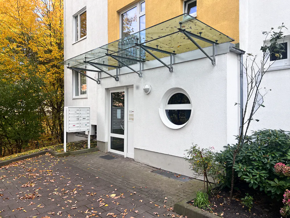 Apartment building entrance with a glass awning, white door, and round window. Autumn leaves on the brick walkway.