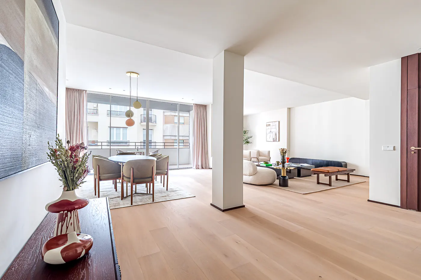 Bright, open-concept apartment with light wood floors. Dining area with round table and chairs, living area with sofas, and a balcony view.