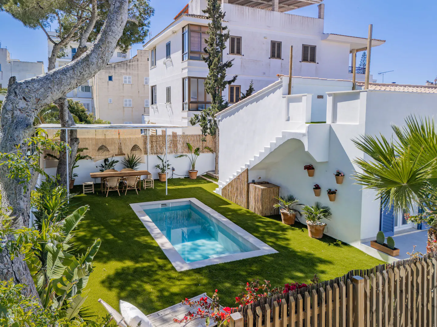 Aerial view of a backyard with a pool, dining area, and white building with stairs. Green grass, plants, and a wooden fence surround the area.