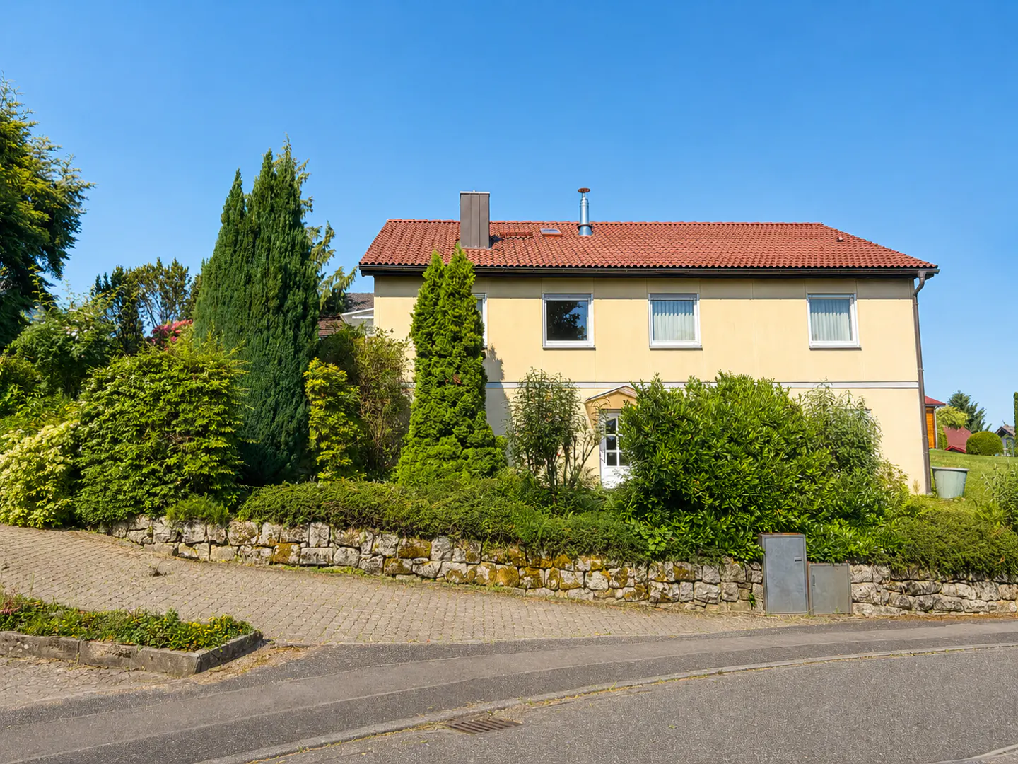 Two-story yellow house with a red tile roof, surrounded by green bushes and trees under a clear blue sky.