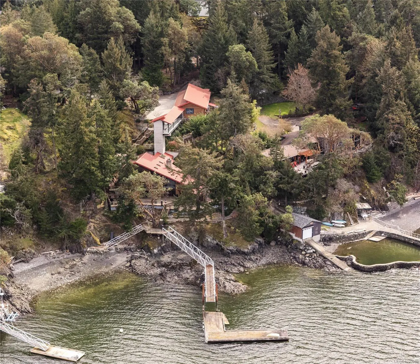 Aerial view of a waterfront property with a red-roofed house, dock, and pool surrounded by lush green trees.