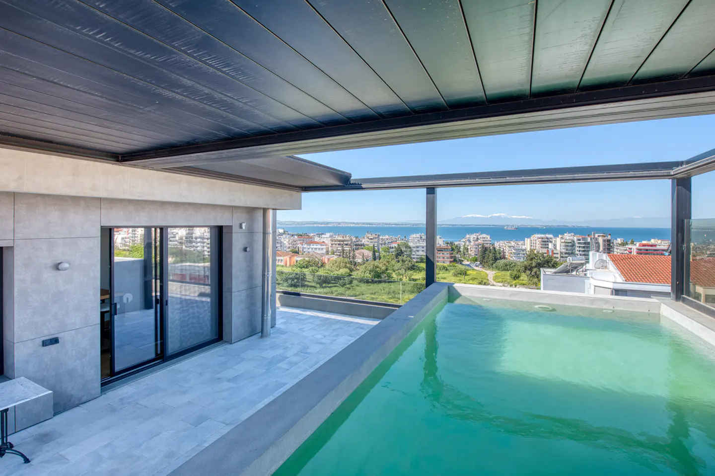 Rooftop view of a pool with city and ocean views. The pool is turquoise, and the city is a mix of buildings and green spaces. The sky is blue.