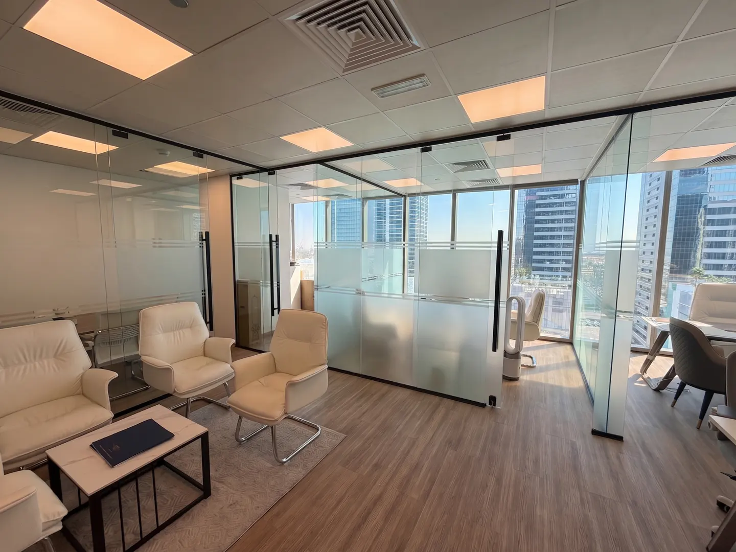 Bright office space with glass-walled rooms, white leather chairs, and a small table with a blue folder. City view visible through windows.