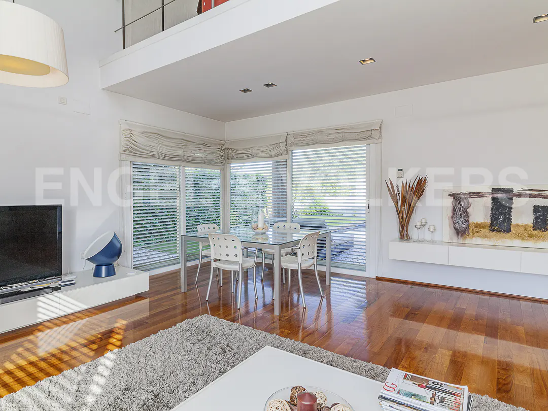 Bright living room with hardwood floors, a glass dining table with white chairs, and large windows with blinds. A gray rug sits in front of a white coffee table.