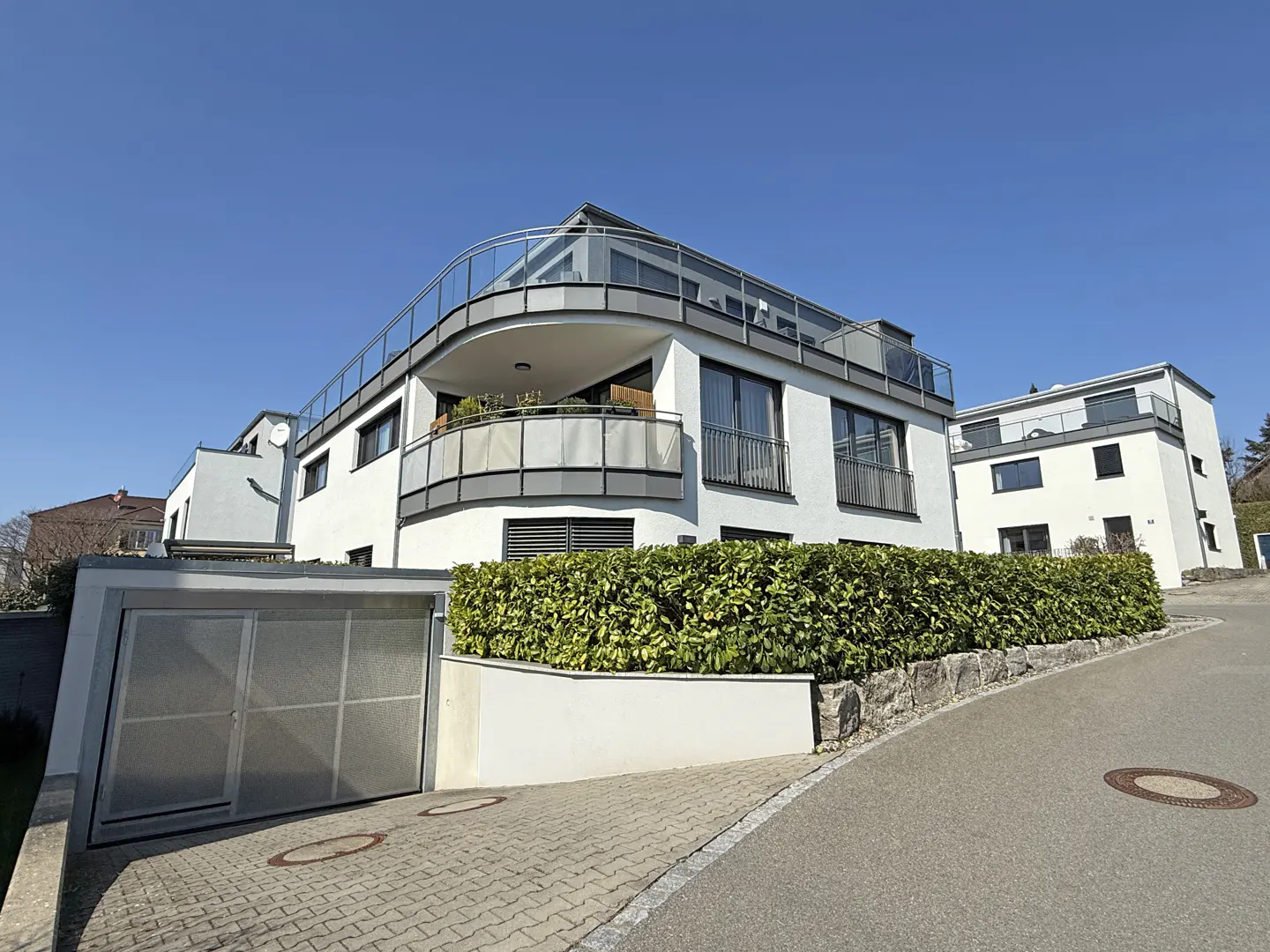 Modern white building with curved balconies and glass railings, set against a clear blue sky. A green hedge lines the property.