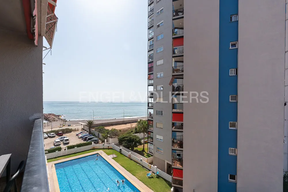 View from a balcony overlooking a blue pool, beach, and ocean, with tall apartment buildings on the right.
