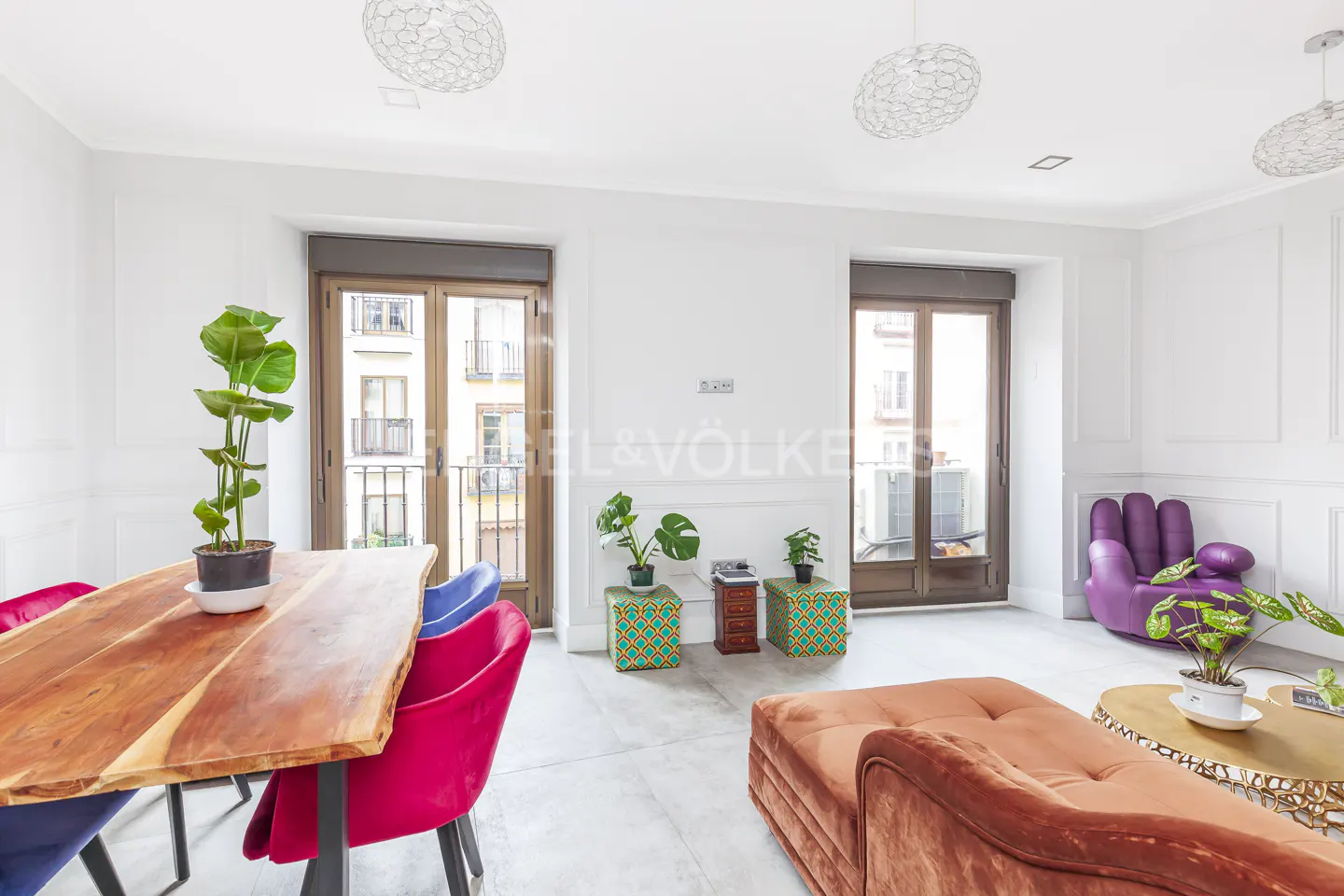 Bright living room with wood table, colorful chairs, brown couch, and purple hand chair. Balconies are visible through the doors.