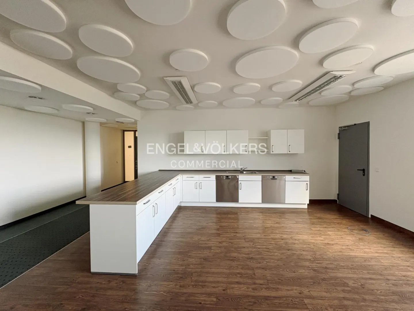 Office break room with wood floors, white cabinets, and a ceiling with round acoustic panels. A gray door is on the right.