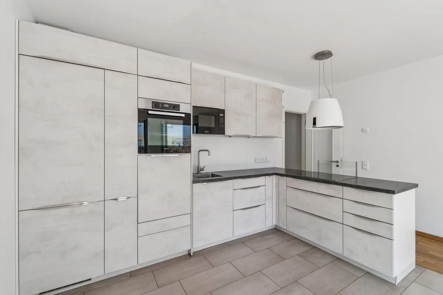 Bright kitchen with gray tile flooring, white cabinets, and black countertops. Stainless steel appliances include an oven and microwave. A white pendant light hangs above the counter.