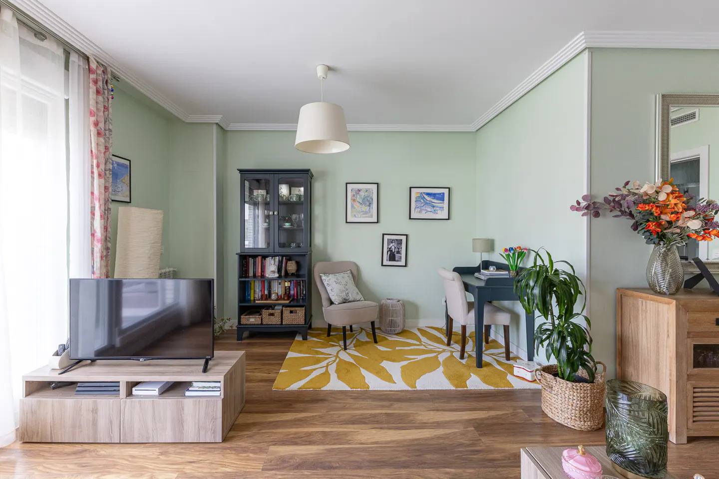 Living room with light green walls, wood floors, a TV on a wood stand, a dark cabinet, a desk, and a yellow leaf-pattern rug.