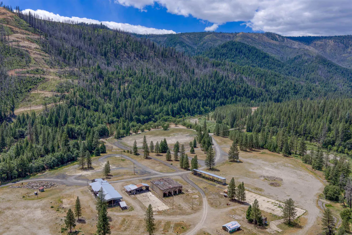 Aerial view of a rural property with several industrial-style buildings, dirt roads, and pine forests against mountains.