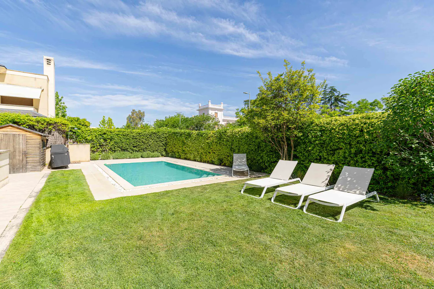 Backyard with a pool, four white lounge chairs on the grass, and a tall green hedge.