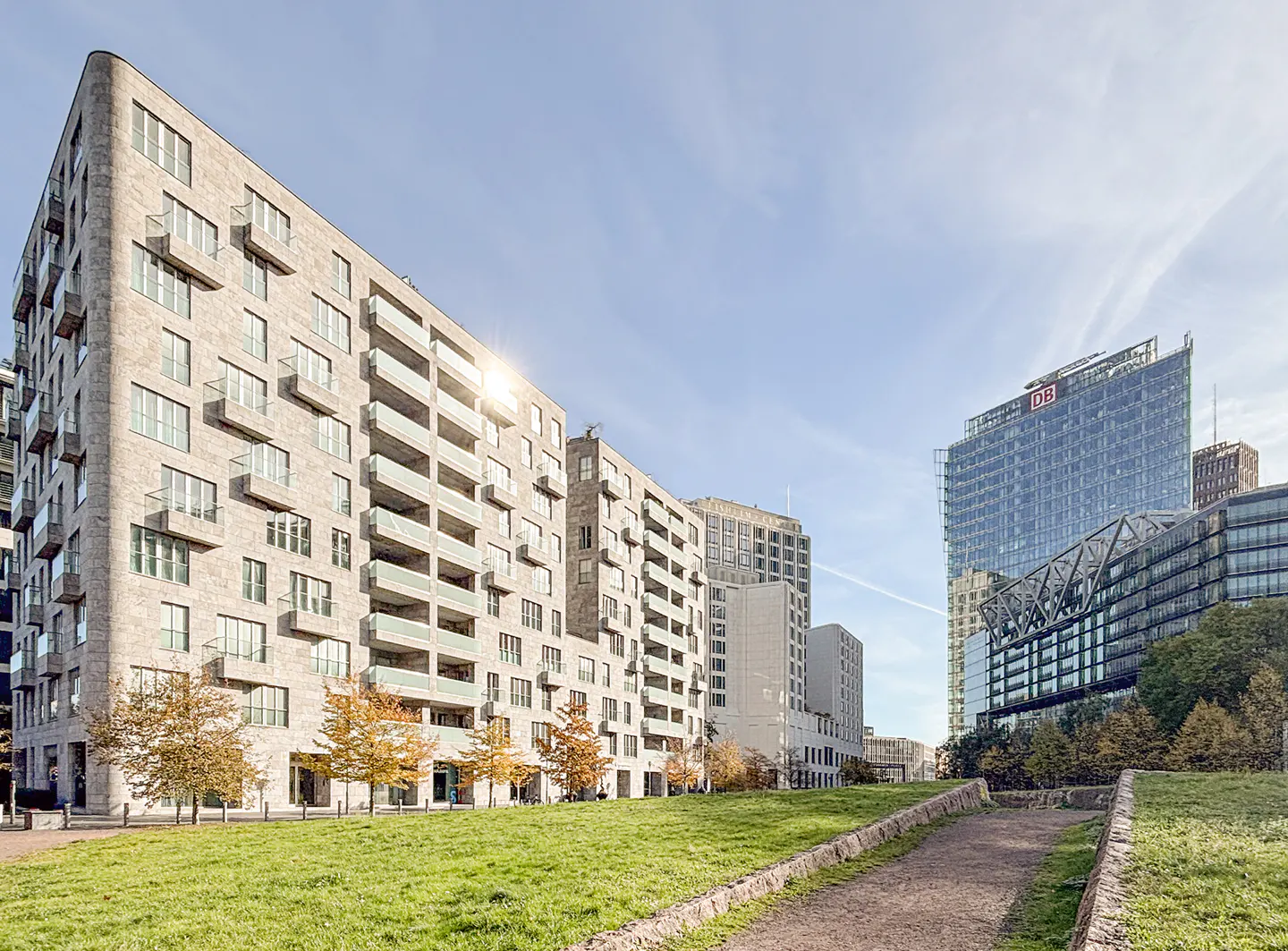 Modern stone apartment buildings with balconies overlook a grassy park with trees under a clear blue sky.