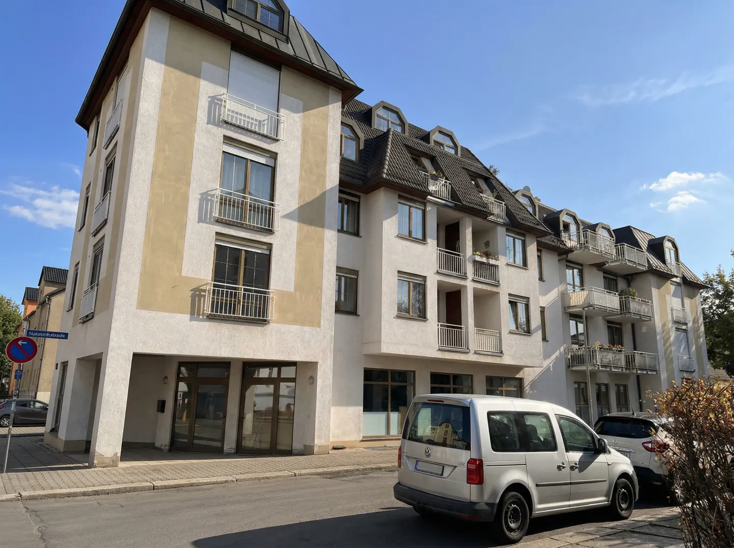 Exterior view of a multi-story apartment building with white and beige facade, balconies, and parked cars on the street.