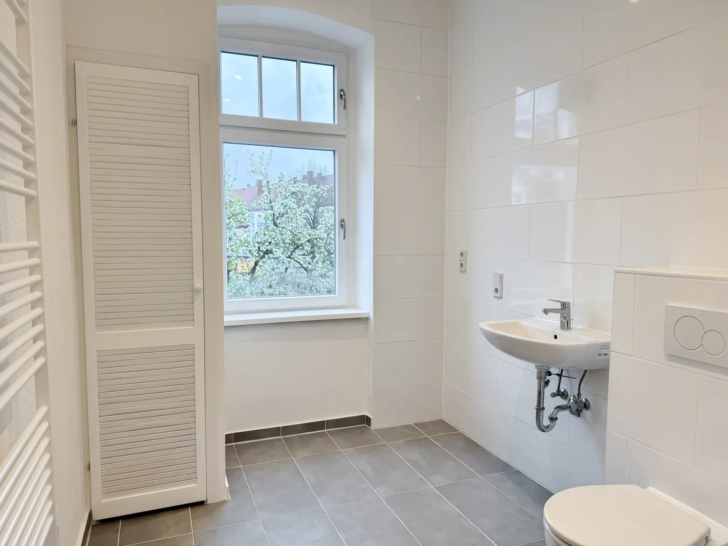 Bright, modern bathroom with white walls, gray tile floor, and a window showing a tree. A white sink and toilet are visible.
