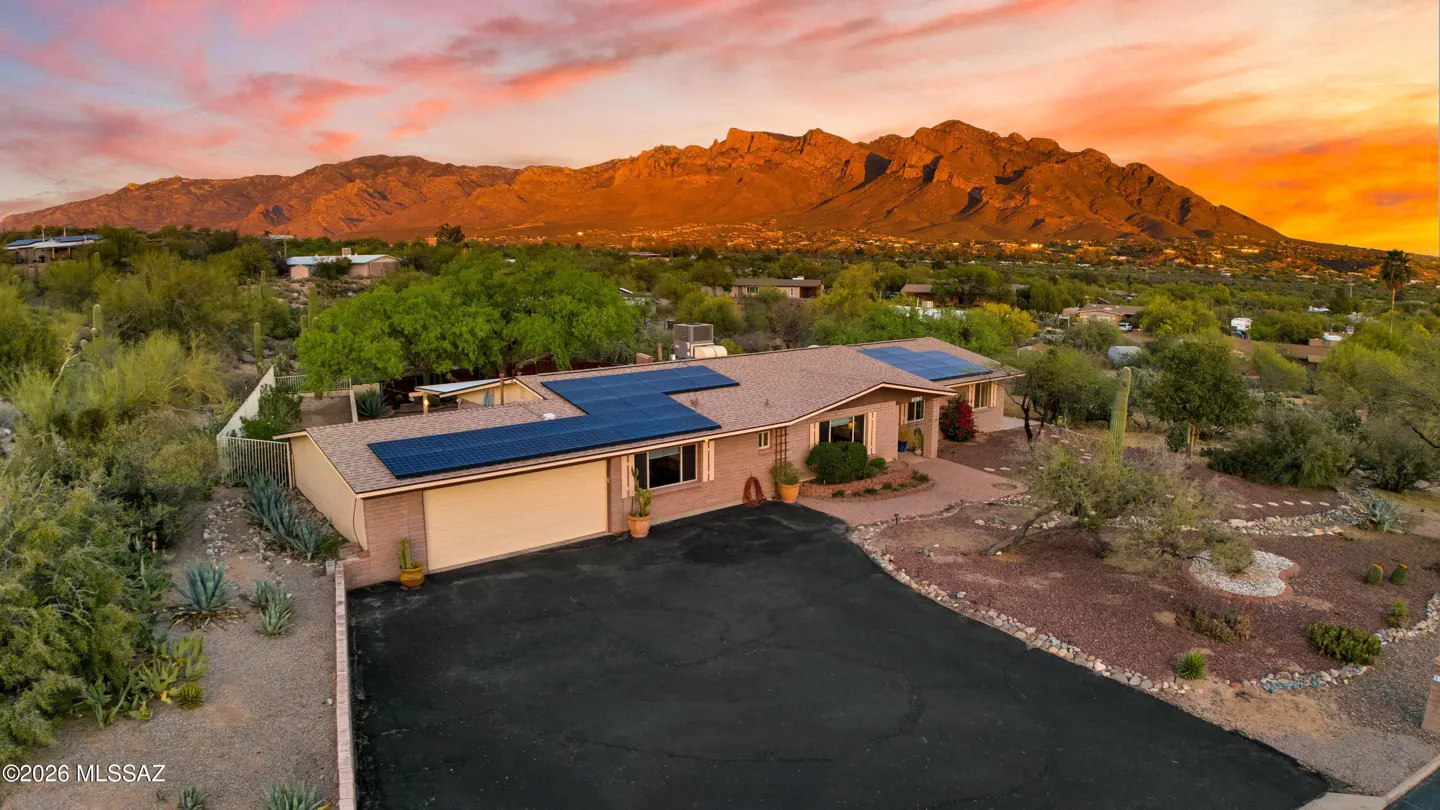 Aerial view of a tan brick house with solar panels, a black driveway, and mountains in the background at sunset.