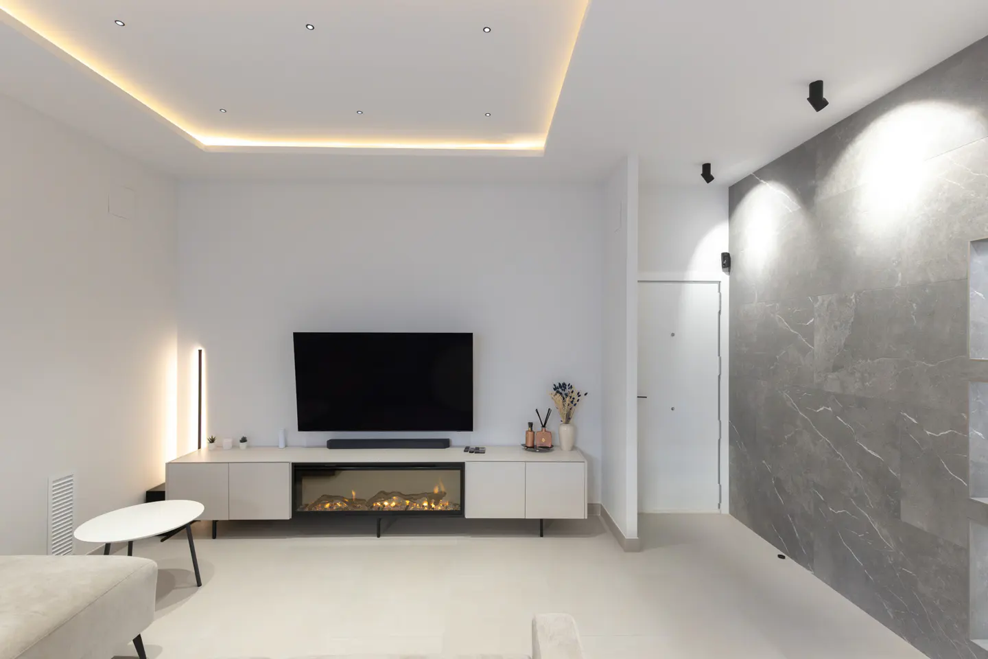 Bright living room with a large TV above a fireplace, a gray marble wall, and recessed lighting.